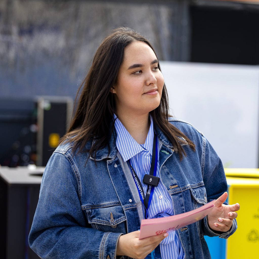 A woman with long dark hair wearing a denim jacket and a striped shirt, holding a pink document and looking thoughtfully to the side.